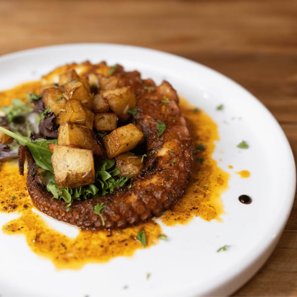 Grilled octopus with crispy potatoes and greens, drizzled with a rich sauce on a white plate.