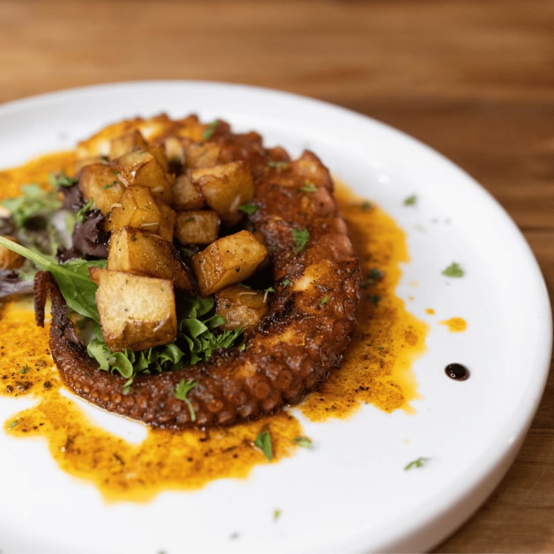 Grilled octopus with crispy potatoes and greens, drizzled with a rich sauce on a white plate.