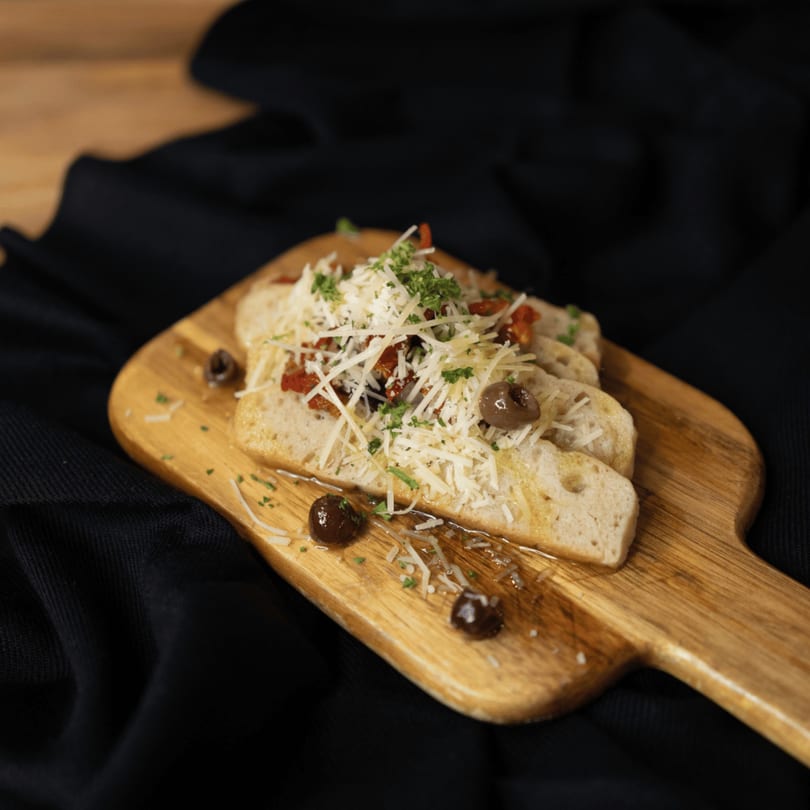 Italian bruschetta with shredded cheese, sun-dried tomatoes, parsley, and olives on a wooden board.